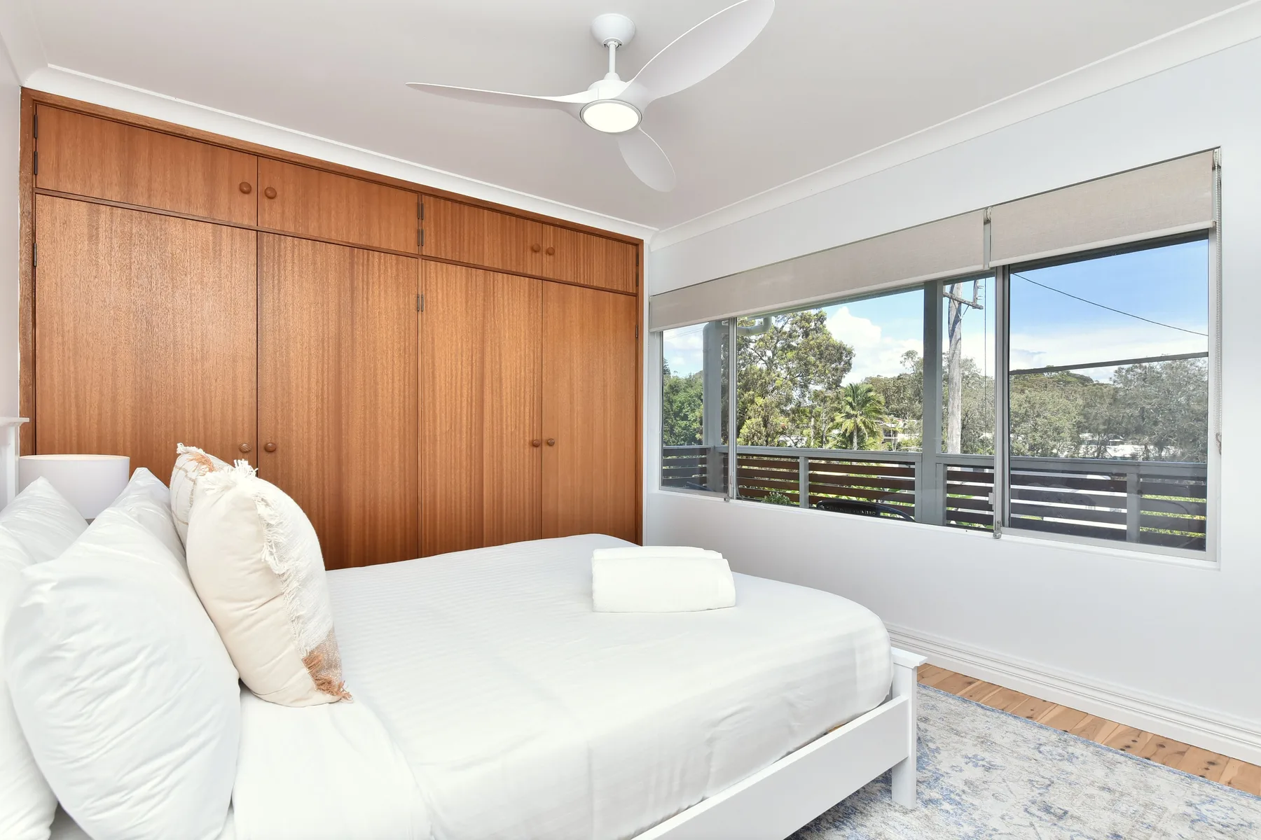 A bright bedroom with a white bed beside large windows overlooking trees.
