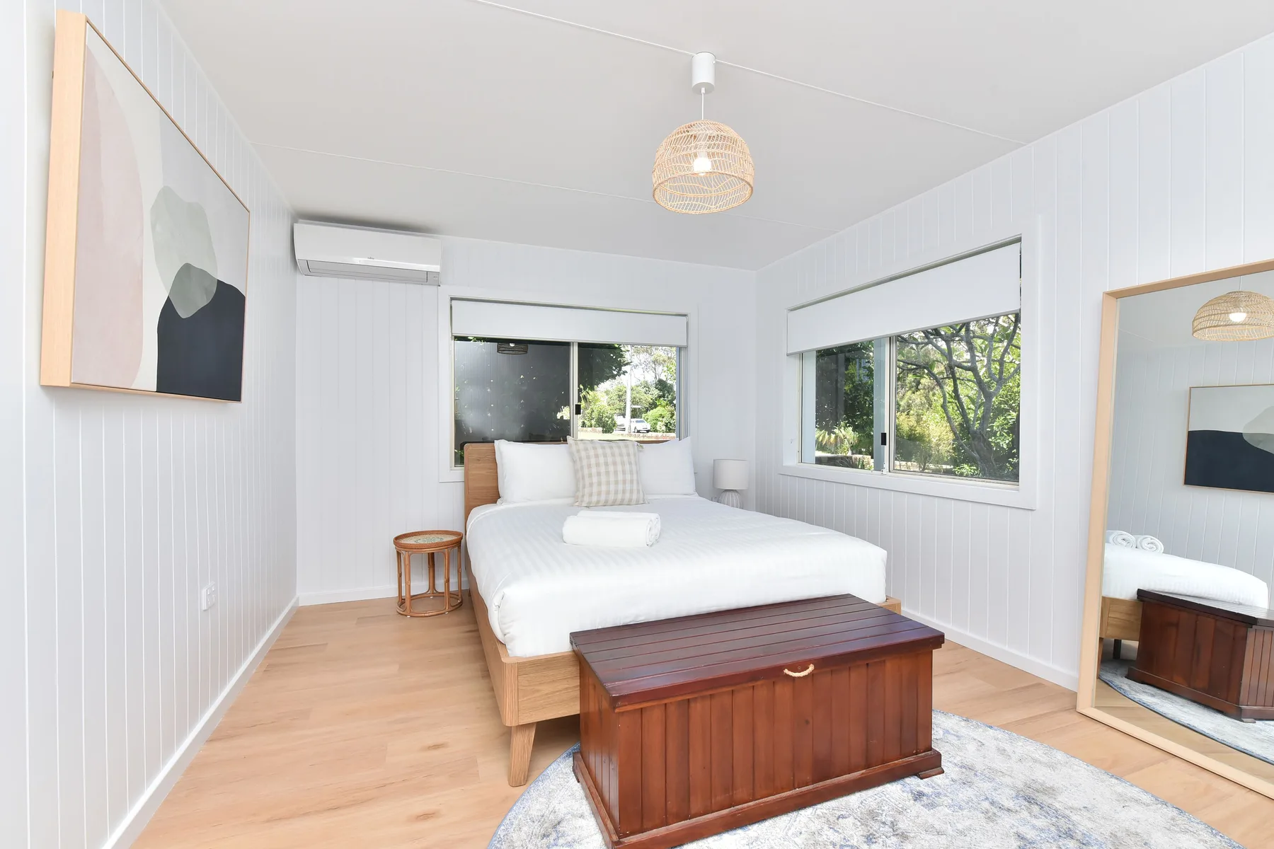 A bright bedroom with a bed, wooden chest, and windows overlooking trees.