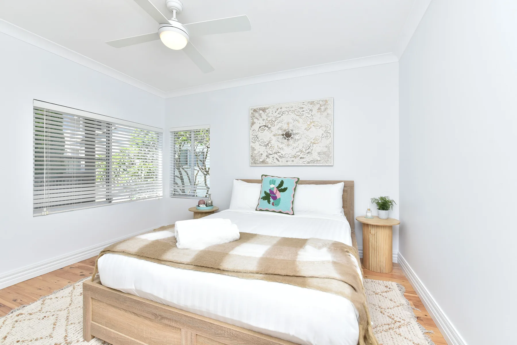 A bright bedroom with a neatly made bed and windows with blinds.