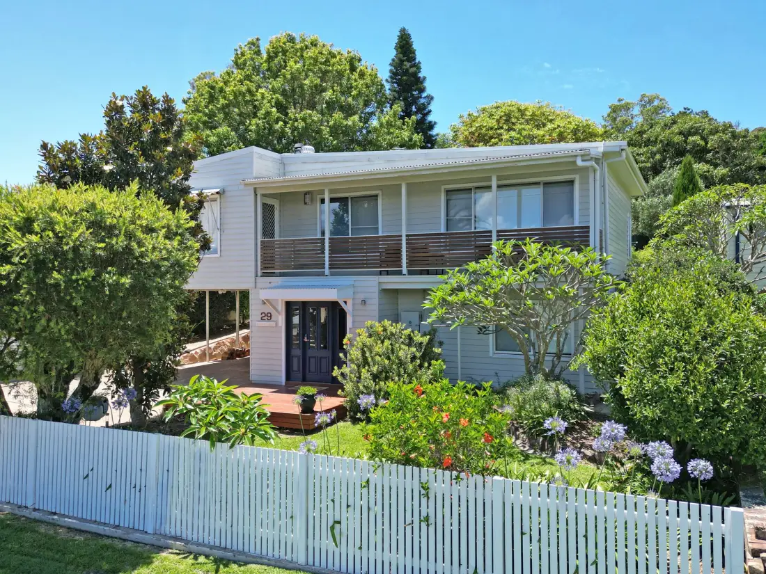 A two-story house with a balcony stands behind a white picket fence and leafy garden.