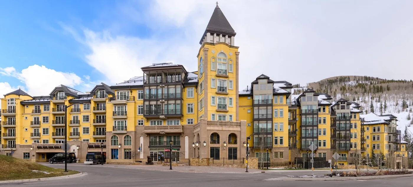 Un gran edificio amarillo de varios pisos con una torre se encuentra junto a una colina nevada.