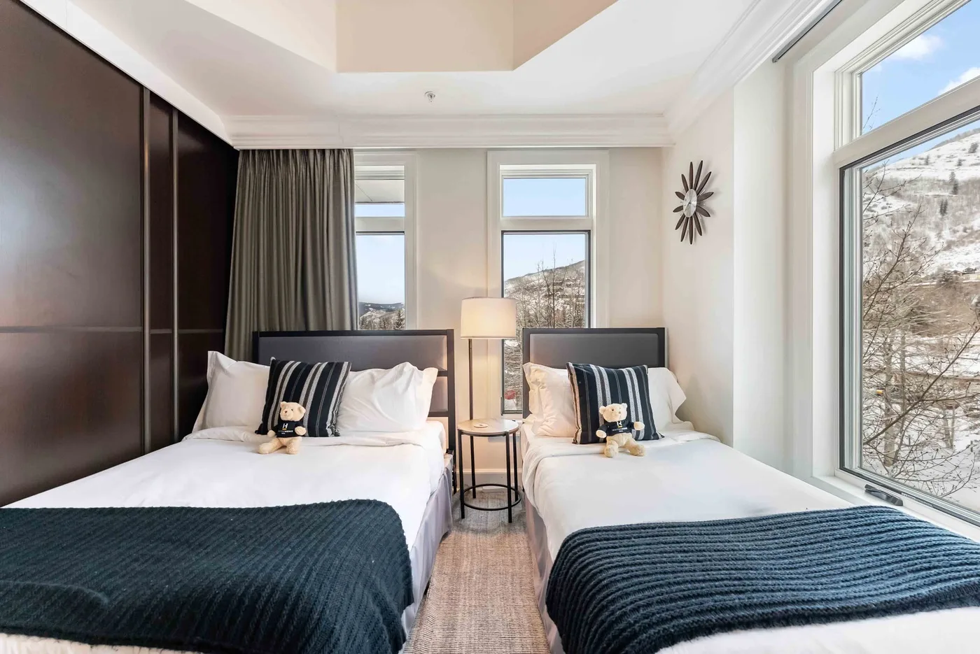 Dormitorio luminoso con dos camas, una mesa auxiliar y grandes ventanas.