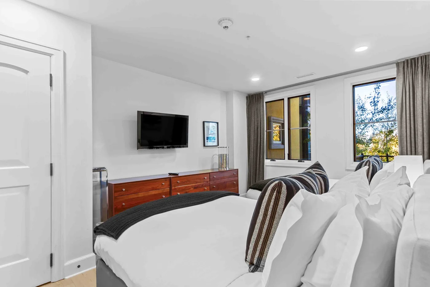 Un dormitorio luminoso con un televisor sobre una cómoda de madera y camas junto a las ventanas.