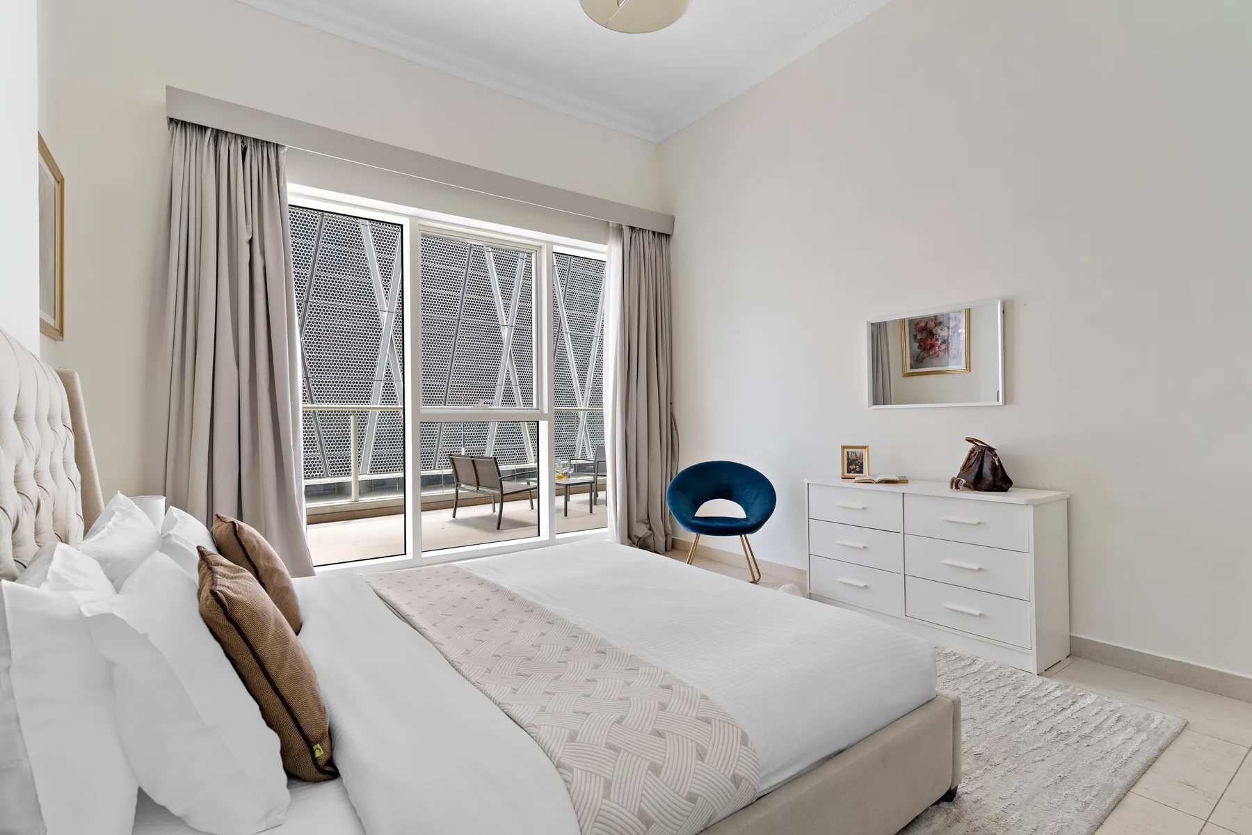 Un dormitorio luminoso con una cama junto a grandes ventanales y una cómoda.