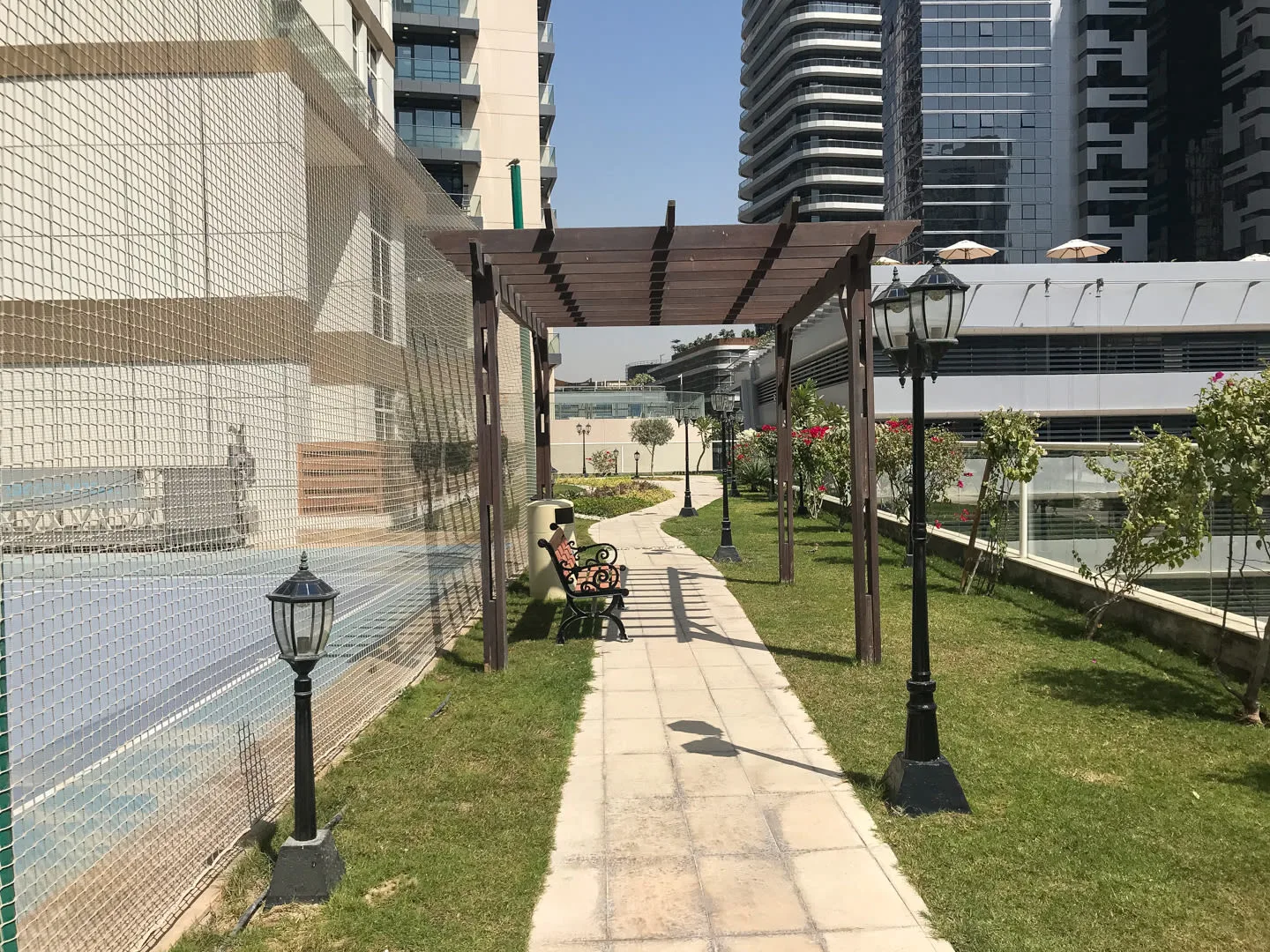 Un sendero pavimentado atraviesa un jardín bajo una pérgola de madera junto a una piscina cercada.