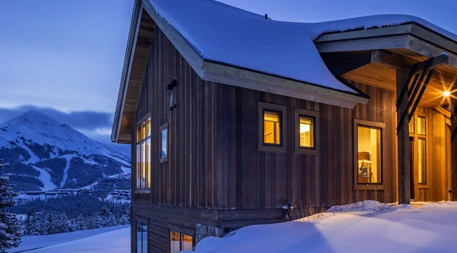 Una cabaña de madera cubierta de nieve brilla con luz cálida frente a montañas al anochecer.