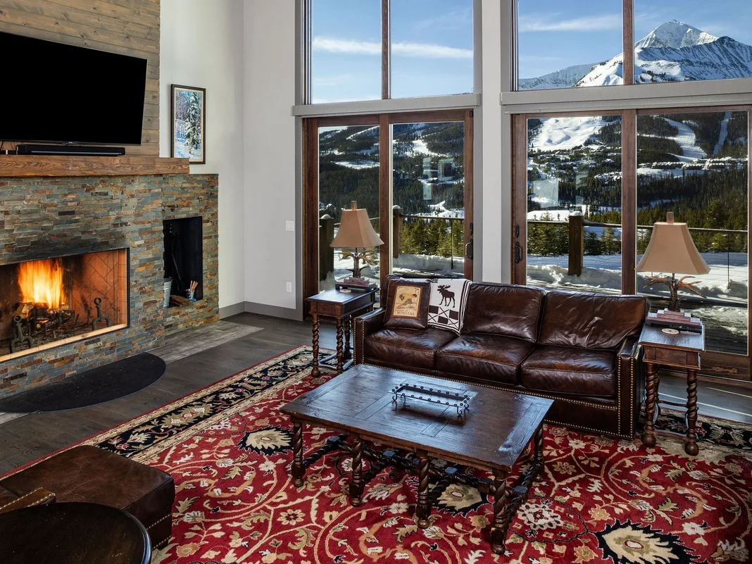 Una sala de estar con sofá de cuero, chimenea y vistas a las montañas por grandes ventanas.