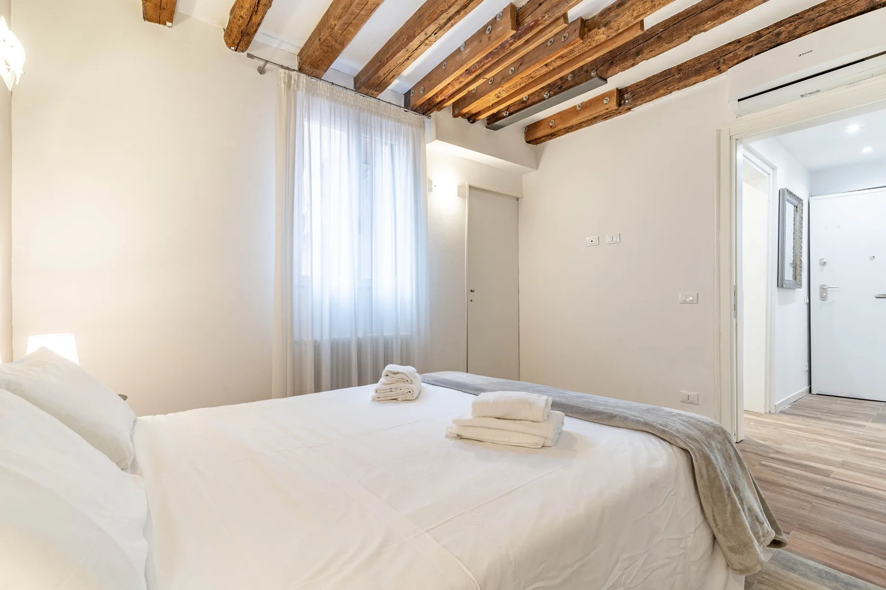 A bright bedroom with a neatly made bed and exposed wooden ceiling beams.