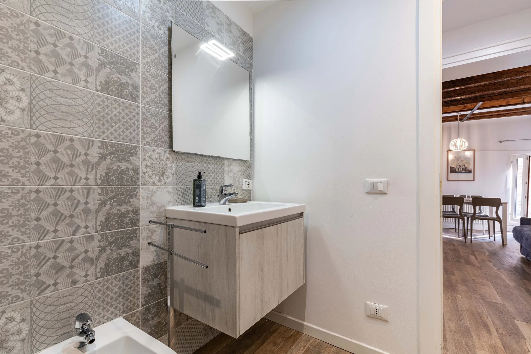 A bathroom vanity and mirror stand beside a wall covered in patterned tiles.