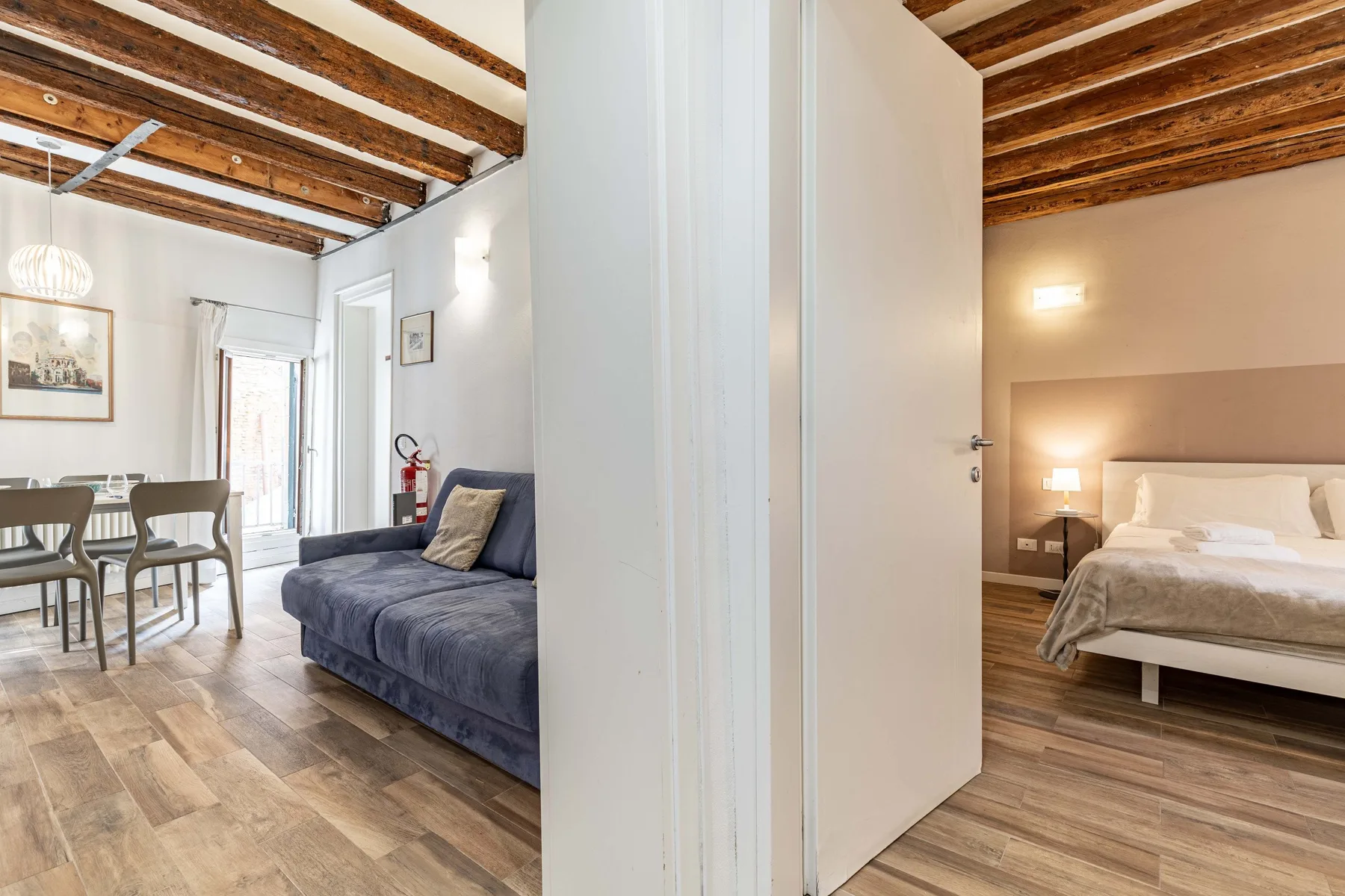 An open doorway separates a bedroom with a bed from a living area with seating and a dining table.