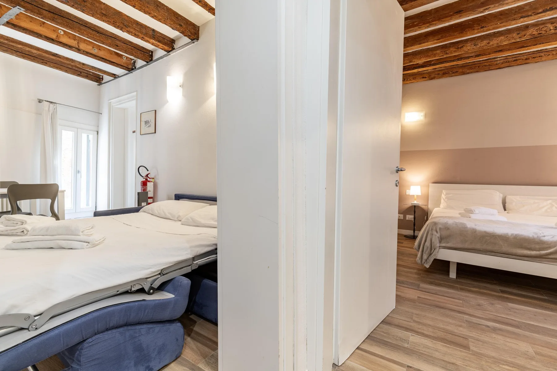 A bedroom with white bedding, exposed wooden ceiling beams, and a light wood floor.
