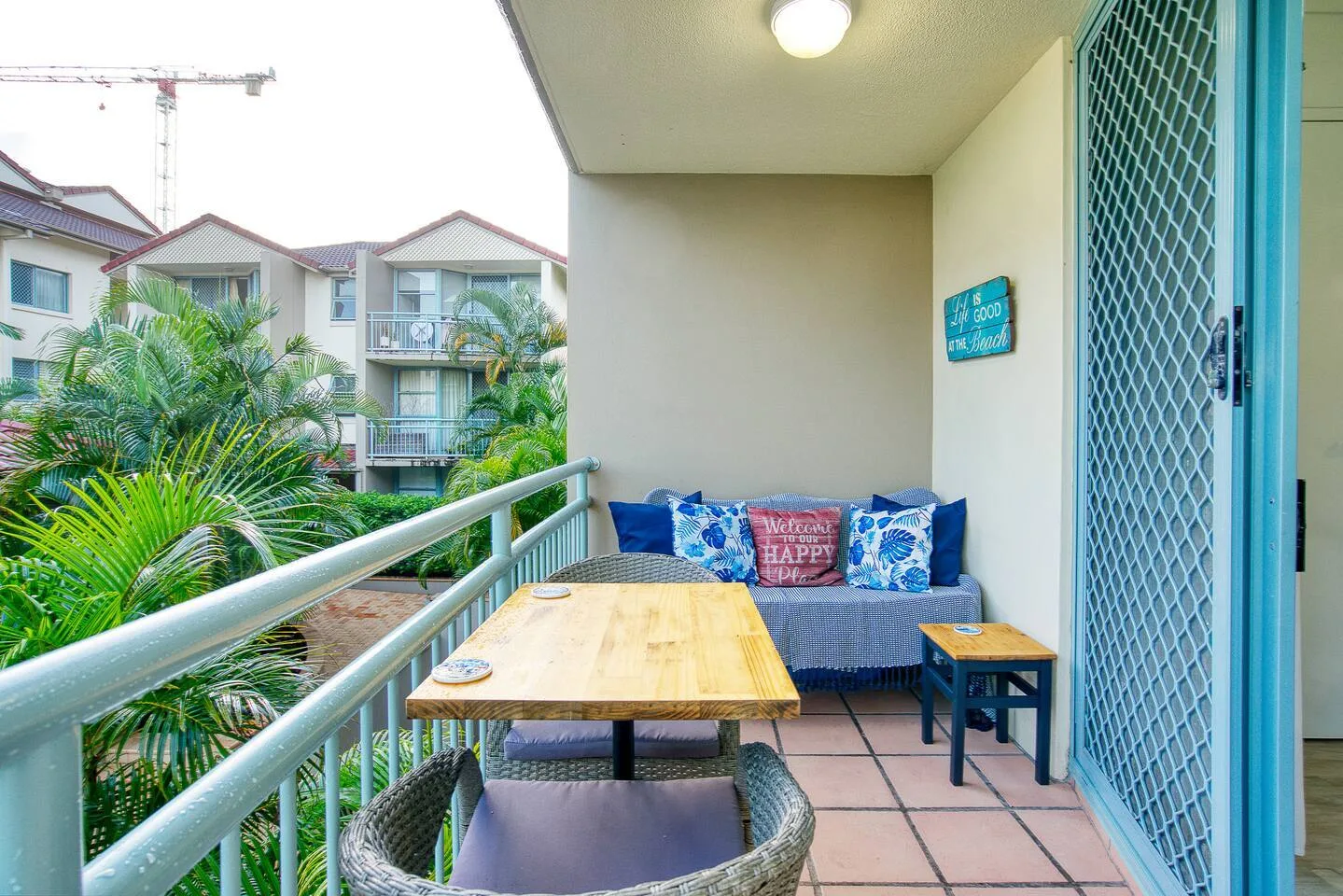 Un balcón con una mesa y cojines azules da a palmeras y edificios cercanos.