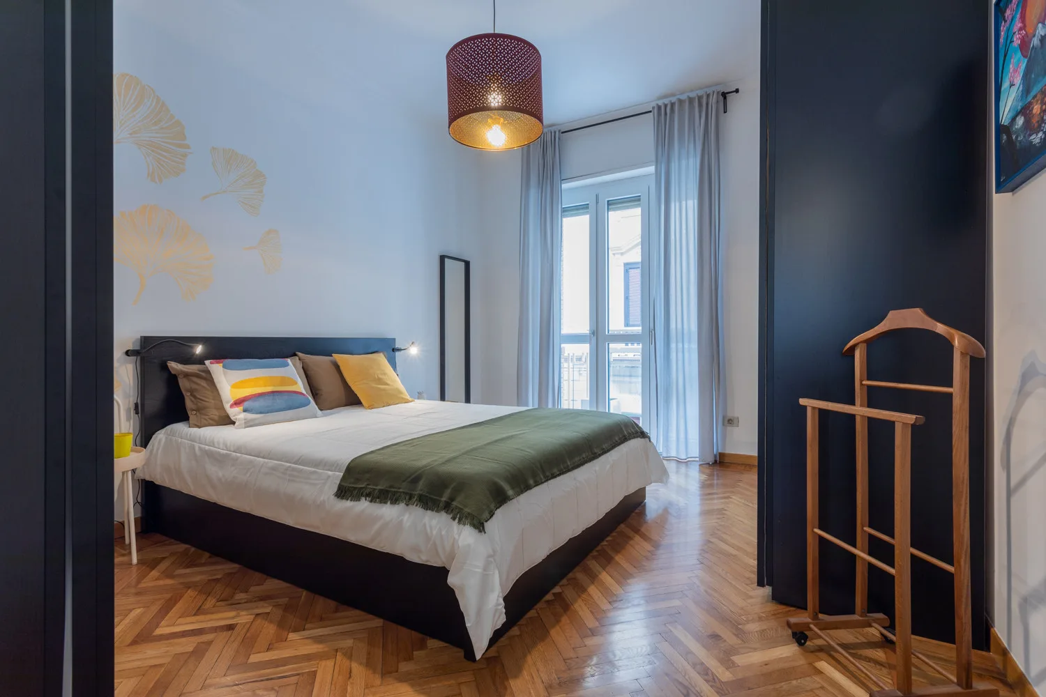 Un dormitorio con una cama, suelo de madera estampado y una ventana alta con cortinas.