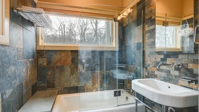 Un baño con bañera, panel de ducha de vidrio y ventanas grandes.