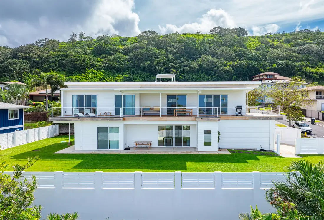 A modern white house overlooks a green lawn, with a forested hillside behind it.