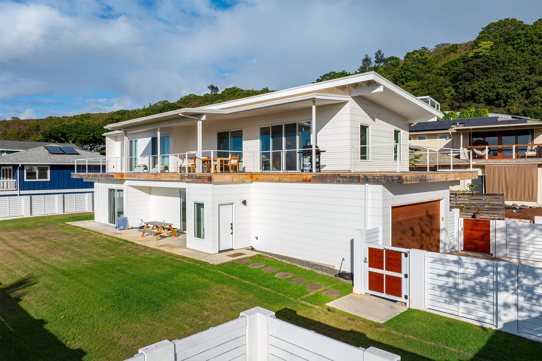 A modern white house with a broad deck overlooks a green lawn.