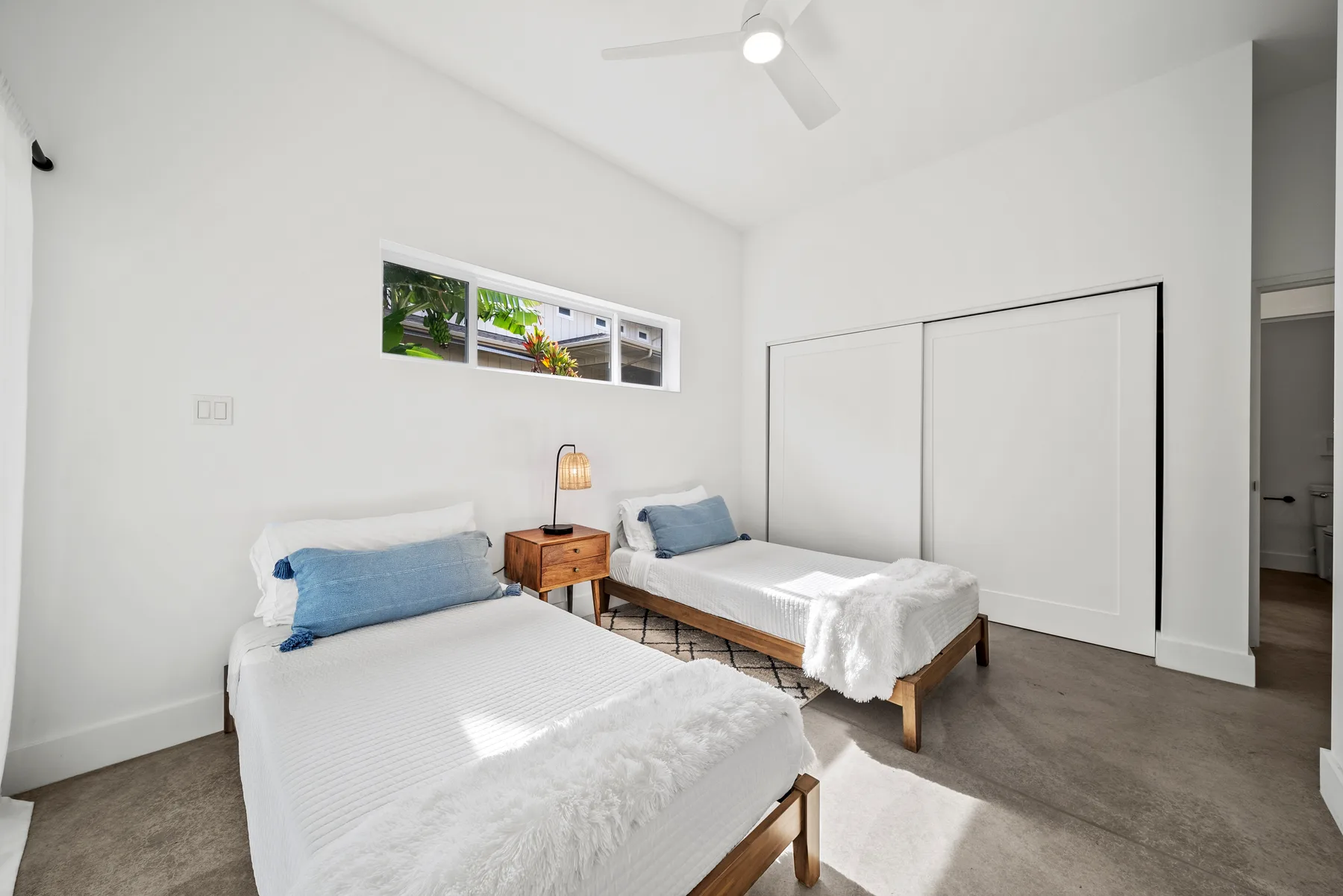 A bright bedroom with two beds, a small bedside table, and a ceiling fan.