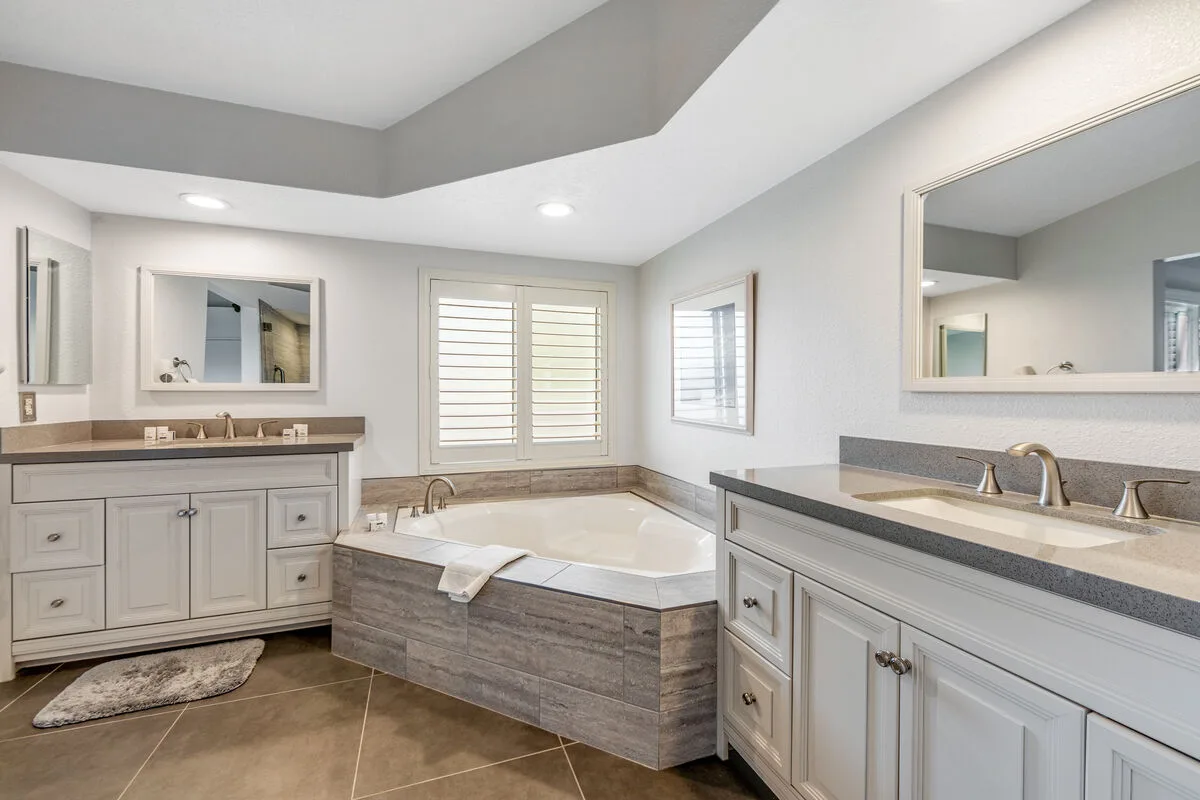 Un baño amplio con una bañera en esquina, muebles de lavabo y ventanas con contraventanas.