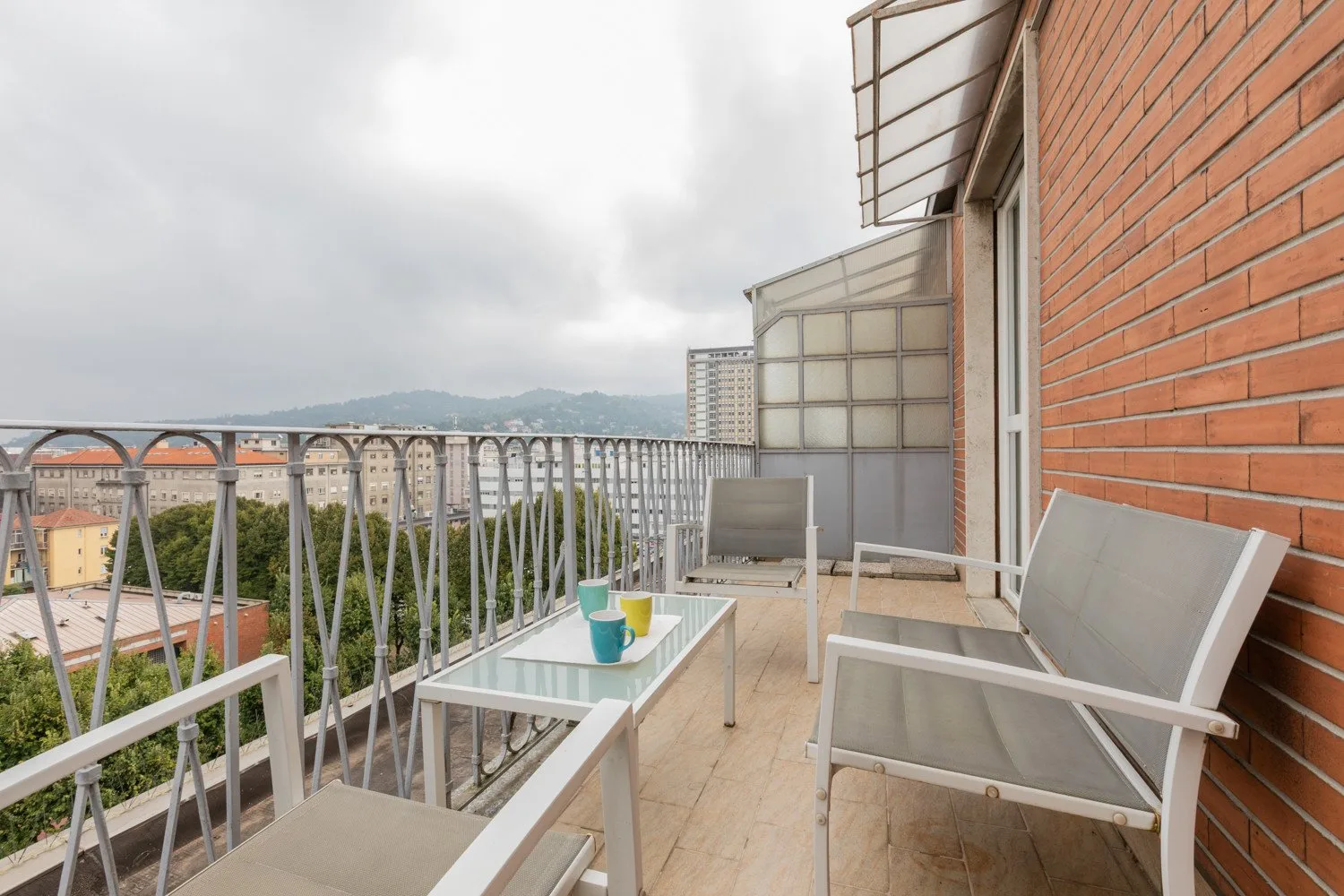 Una zona de estar en el balcón con una mesa de vidrio da a tejados y colinas cercanas.