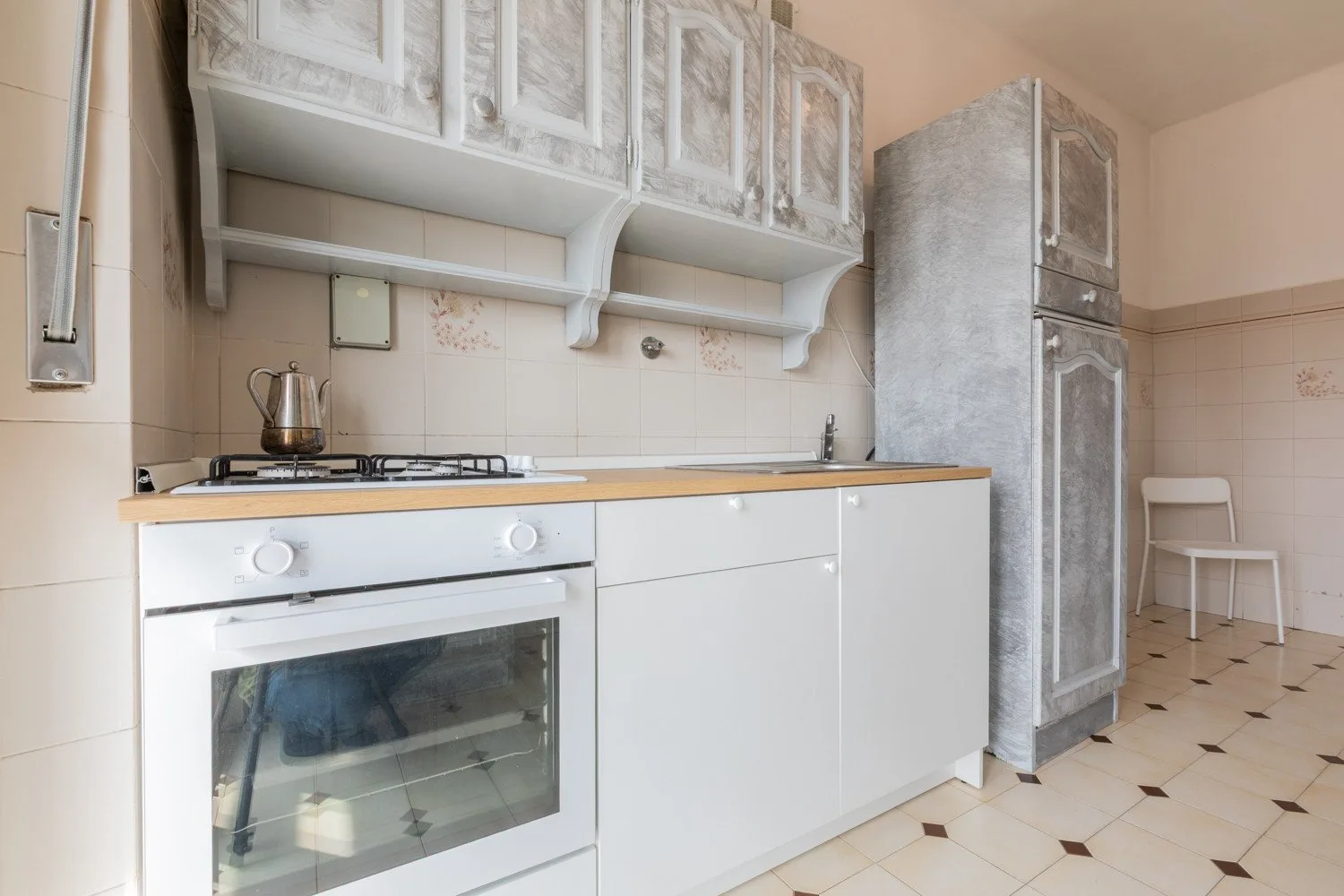 Una cocina alicatada con armarios blancos, una placa de cocina y un refrigerador alto.
