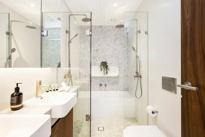 Un baño luminoso con ducha de vidrio, lavabo blanco y detalles de madera.