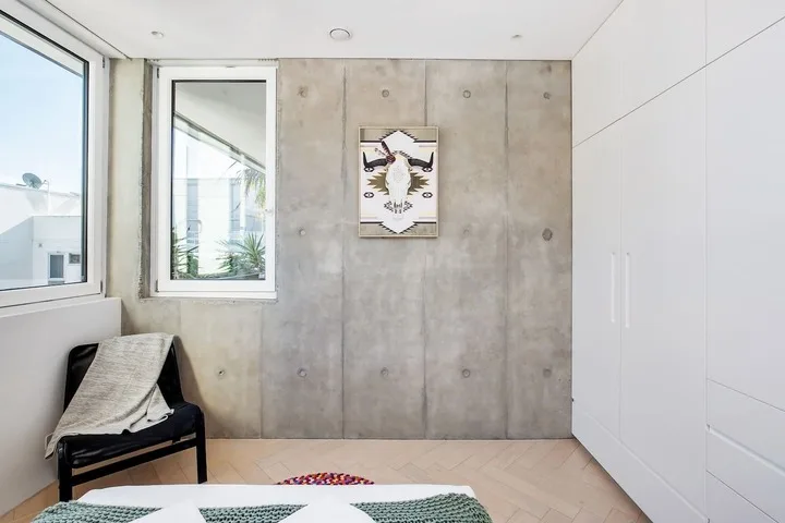 Un dormitorio con una cama, una silla y una ventana grande junto a una pared de concreto.