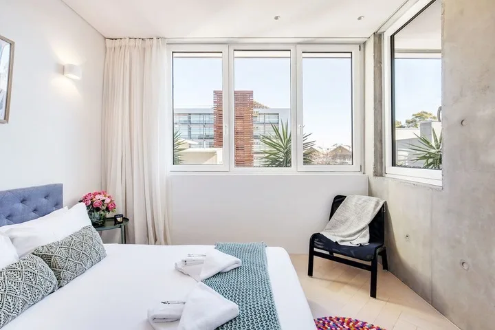 Un dormitorio luminoso con una cama, una silla y ventanas grandes.