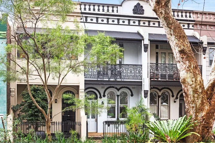 Edificio blanco de dos plantas con balcones ornamentados y árboles delante.