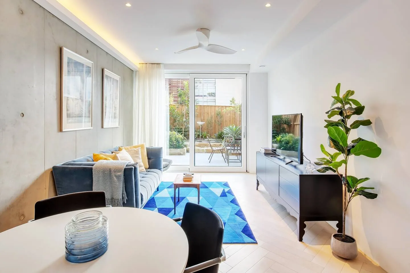 Una sala luminosa con asientos, una alfombra azul y una puerta de vidrio al patio.