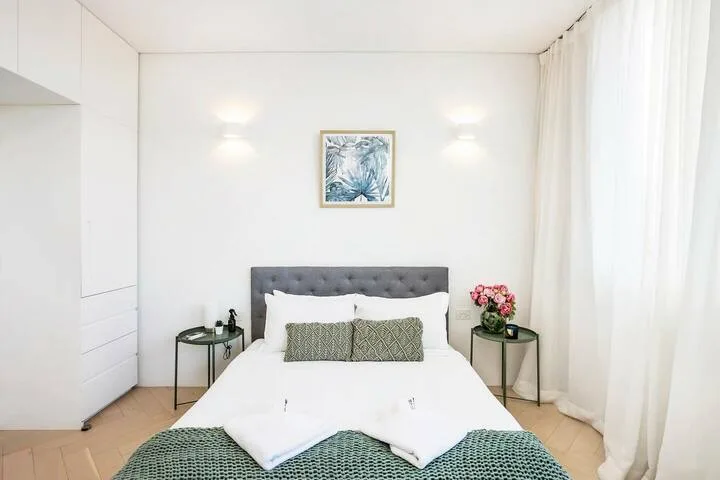 Un dormitorio luminoso con cabecero tapizado gris y ropa de cama blanca.