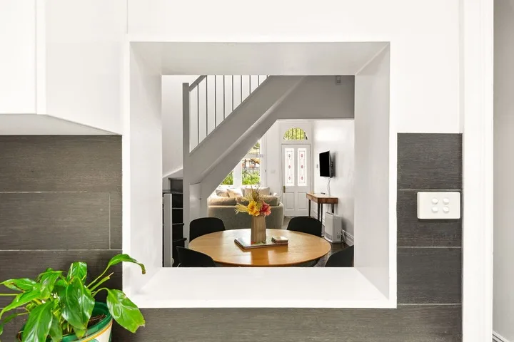 Un comedor está bajo una escalera, con una mesa redonda y un jarrón con flores.