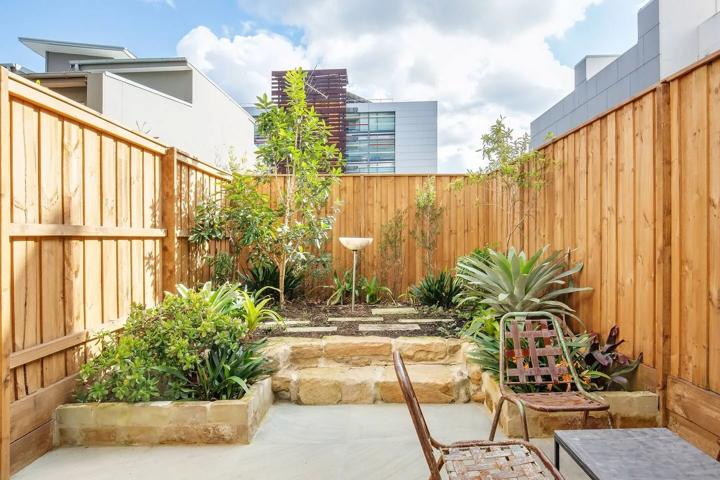 Un patio ajardinado tiene jardineras elevadas de piedra, cercas de madera y asientos al aire libre.