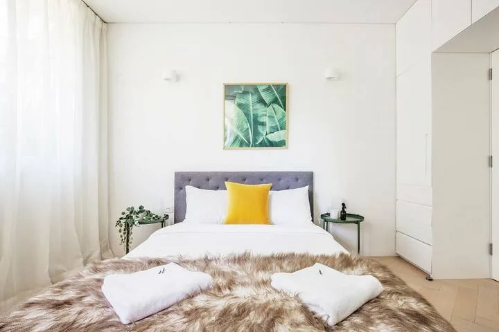 Un dormitorio luminoso con una cama, un cojín amarillo y arte botánico en la pared.