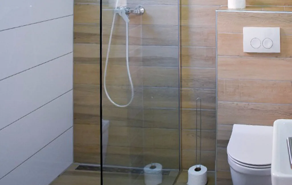 A bathroom with a glass shower, toilet, and wall-mounted flush button.