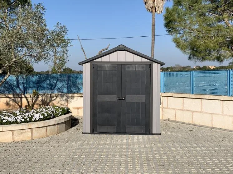 Un pequeño cobertizo de almacenamiento está en un patio pavimentado junto a un arriate.