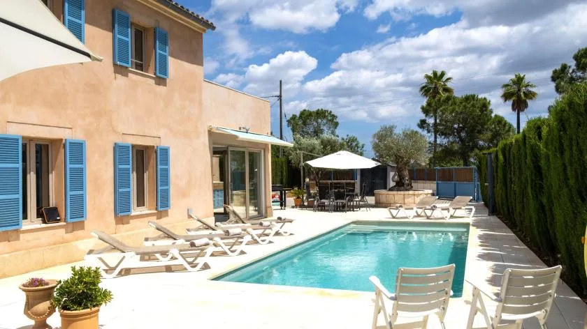 Una piscina junto a una casa color durazno, con asientos de descanso en el patio.