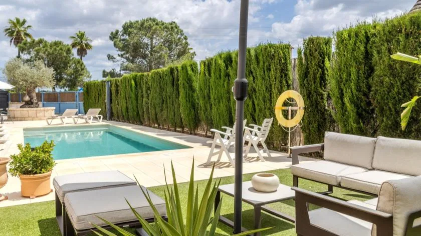 Una piscina al aire libre junto a una zona de estar en el patio, con setos altos alrededor.