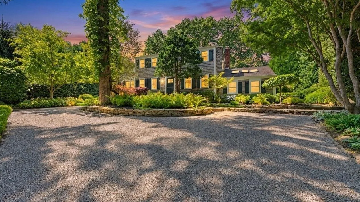 Una casa grande está rodeada de árboles maduros junto a una amplia entrada pavimentada.