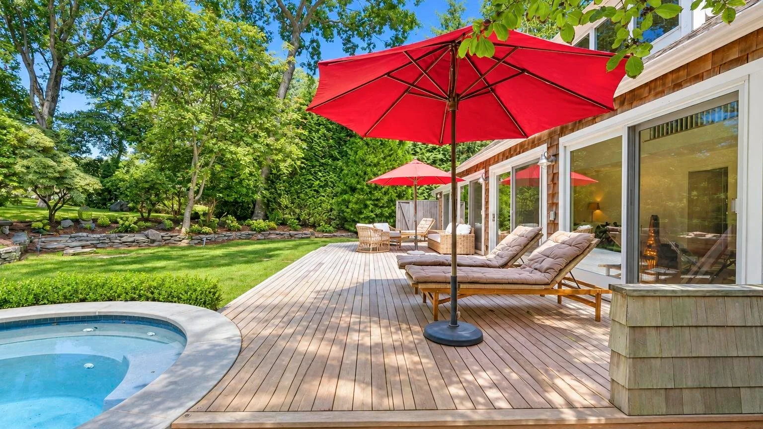 Una terraza de madera junto a una piscina tiene asientos reclinables y una sombrilla roja.