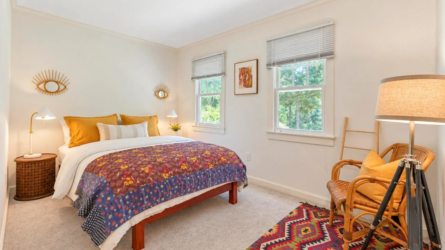 Un dormitorio luminoso con una cama, una colcha colorida y dos ventanas.