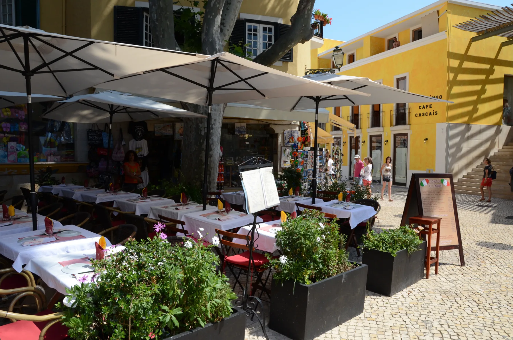 An outdoor café terrace with shaded tables and planters beside a pedestrian street.