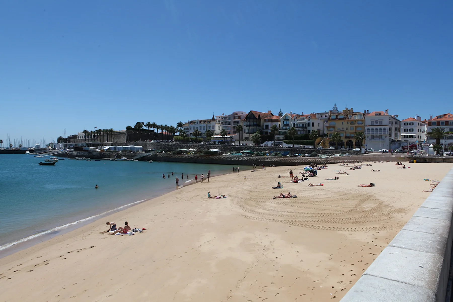 A sandy beach curves along calm blue water, with buildings lining the shore.