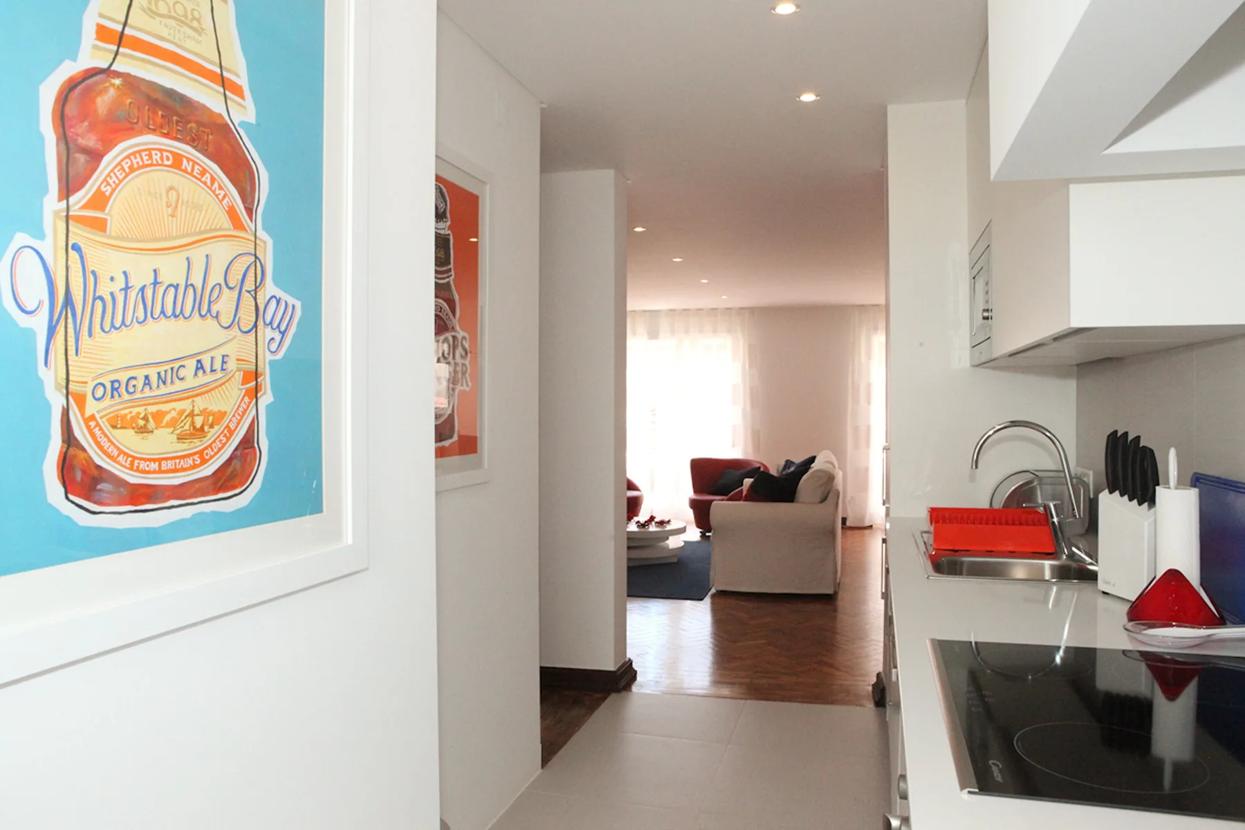 A kitchen corridor opens toward a living area with seating and a bright window.