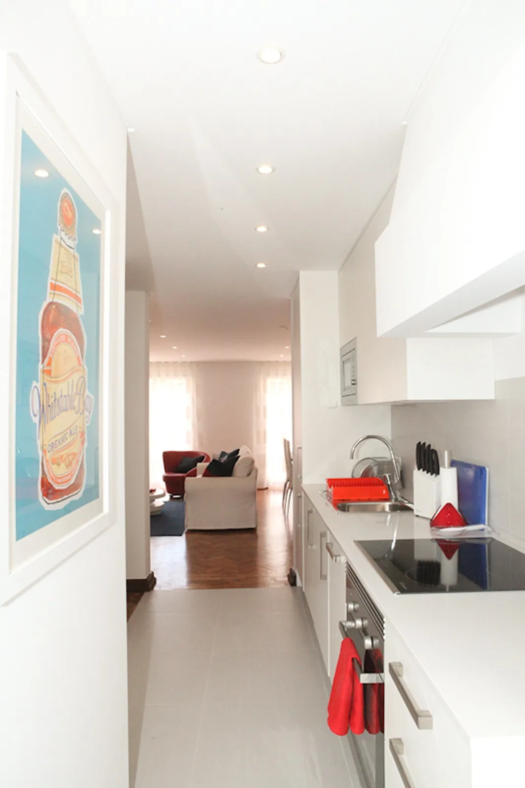 A narrow kitchen opens into a living area with seating and a framed wall print.