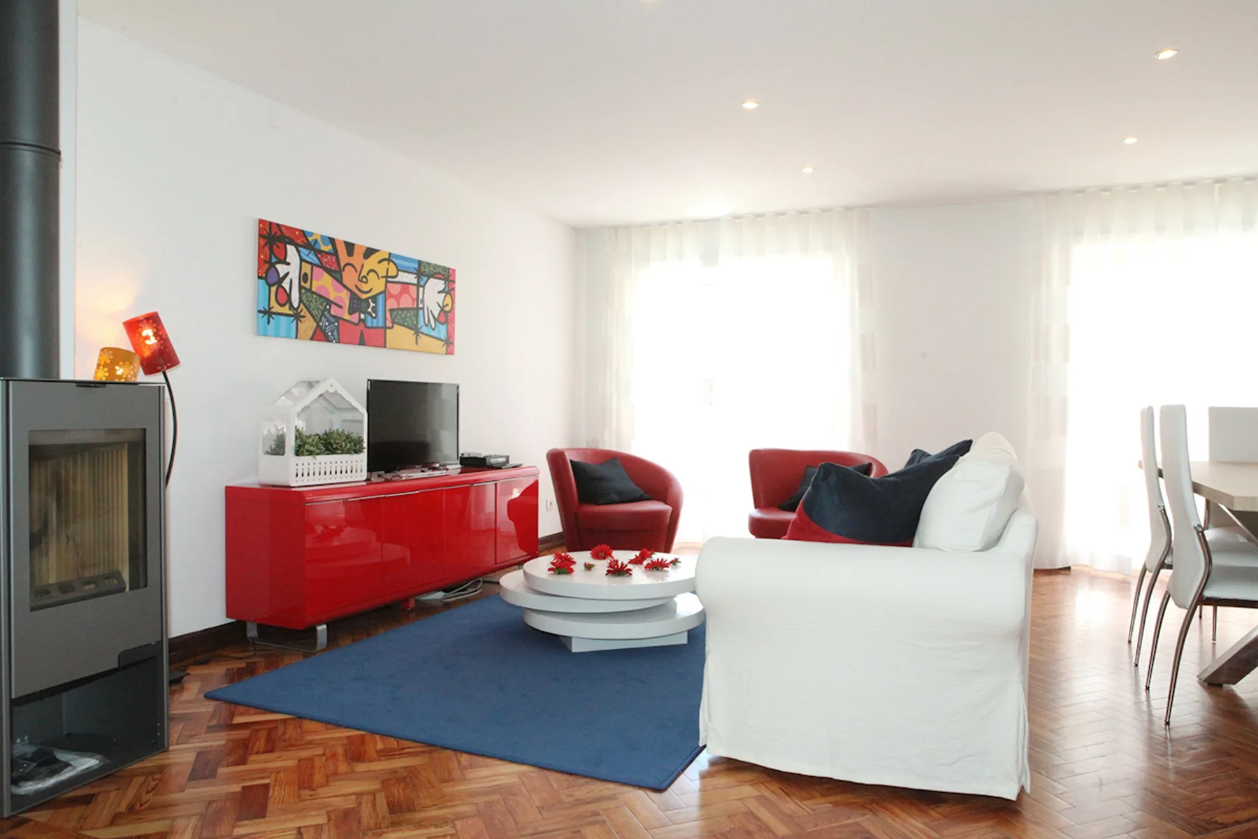 A bright living room with a seating area, red cabinet, and blue rug.