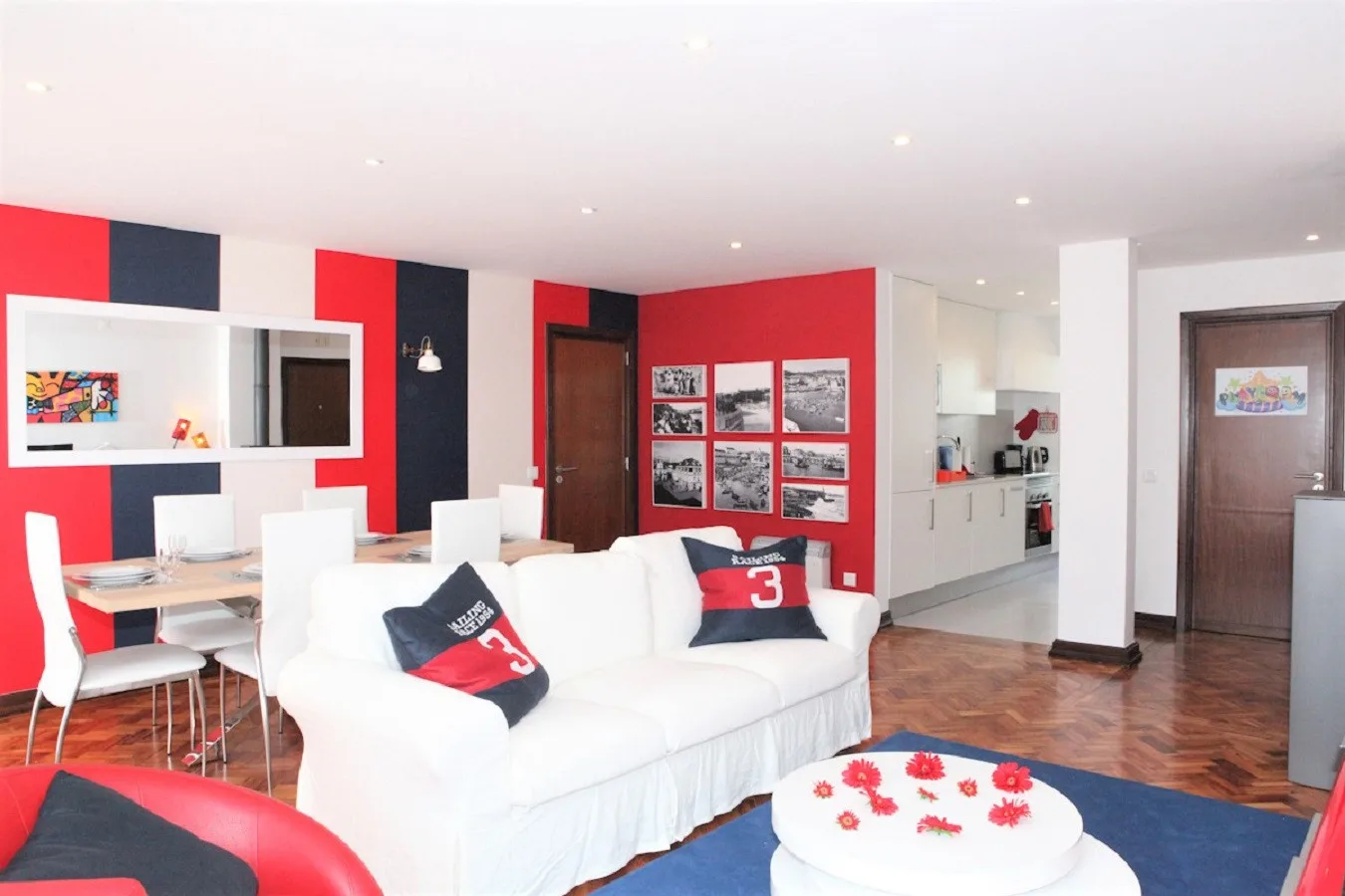 Una sala luminosa con asientos blancos, una pared roja y una cocina contigua.