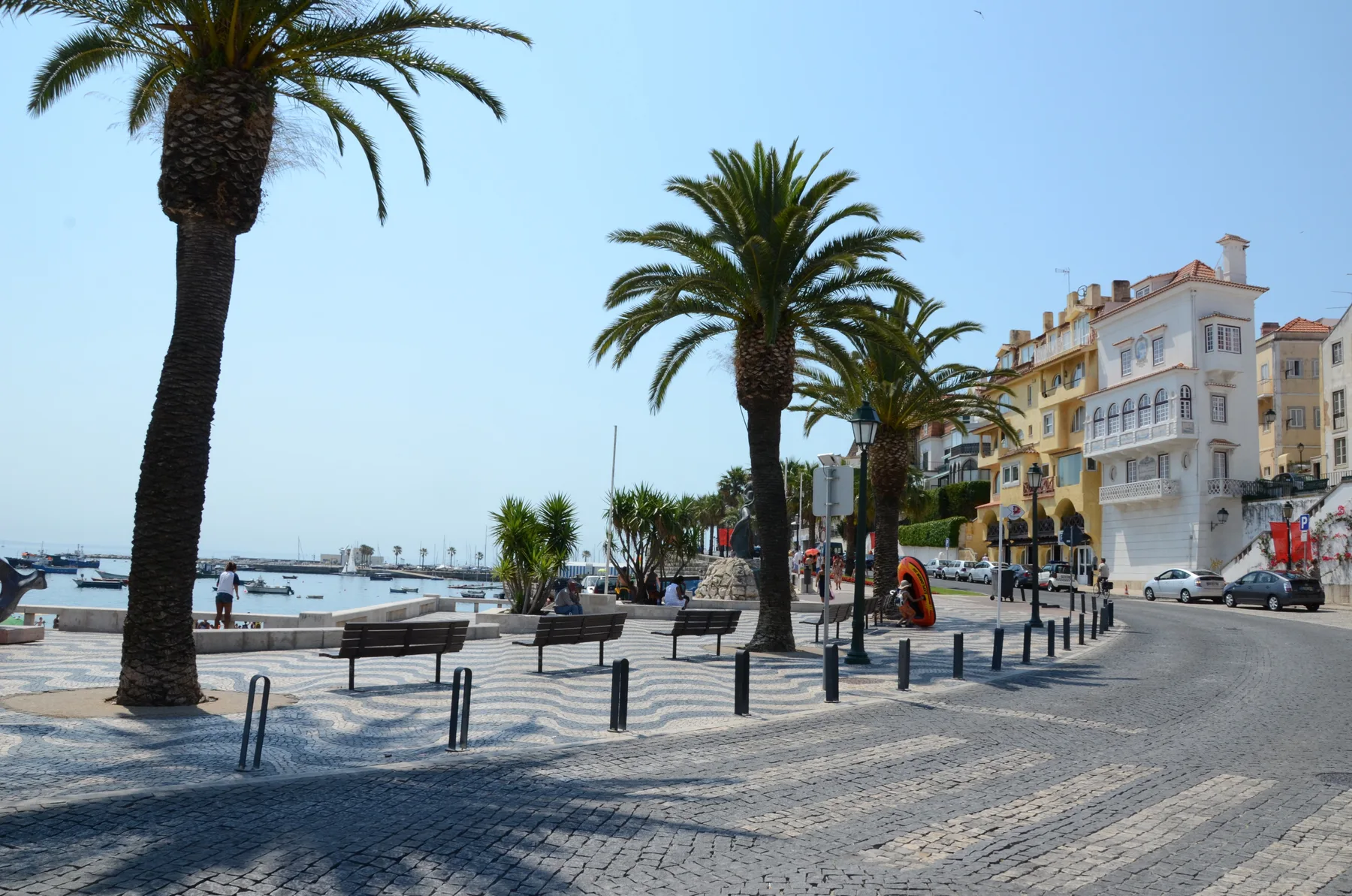 A seaside promenade with palm trees, outdoor seating, and buildings along the road.
