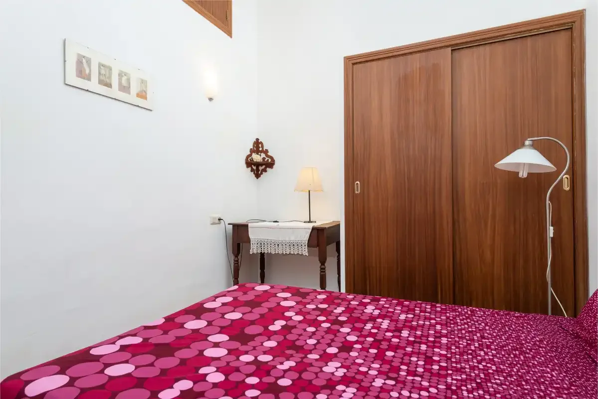 A bedroom with a patterned bedspread, wooden wardrobe, and small table.