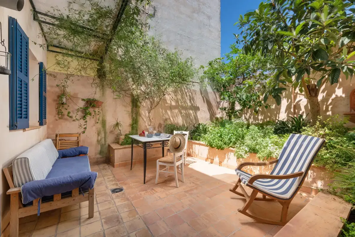 A sunlit outdoor patio with seating, potted plants, and leafy trees.