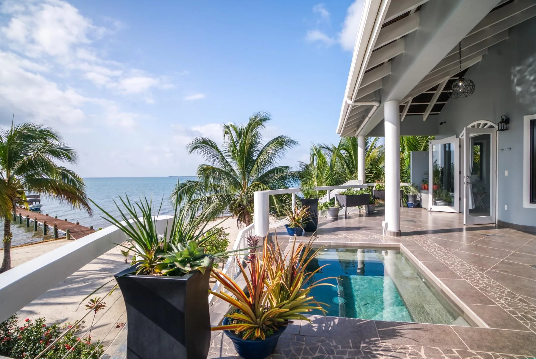 A seaside patio with a small pool, potted plants, and palm trees.
